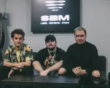 Trzech mężczyzn siedzi na kanapie. W tle logo SBM. Może to scena z teledysku lub sesji nagraniowej piosenki Bedoesa.