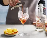 Ein Aperol-Spritz-Cocktail mit Prosecco wird zubereitet. Eiswürfel im Glas, daneben Orangenstücke und eine Flasche Sprudelwasser.