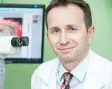 Okulista z uśmiechem przy aparaturze do badań. Na ekranie widok oka, sugerujący diagnostykę przed przeszczepem rogowki.