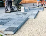 Układanie płyt betonowych na podjeździe – alternatywa dla kostki. Pracownicy układają szare płyty na podsypce żwirowej.