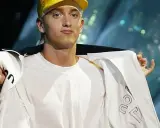 Slim Shady w żółtej czapce i białej bluzie, z gestem dłoni.