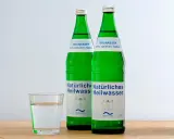 Zwei grüne Flaschen Heilwasser und ein Glas Wasser. Ein Glas Wasser vor dem Schlafengehen trinken kann beim Abnehmen helfen.