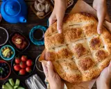 Hände halten ein frisch gebackenes Ramadan Brot, bestreut mit Sesam und Schwarzkümmel. Daneben liegen kleine Schalen mit Oliven, Tomaten und anderen Köstlichkeiten.