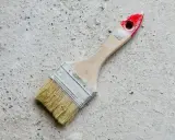 Pędzel do malowania betonu na zewnątrz, z drewnianą rączką i czerwonym zakończeniem, leży na szarej, chropowatej powierzchni.