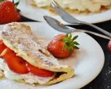 Puszysty omlet z białek, nadziany kremem i truskawkami, posypany cukrem pudrem. Obok leżą świeże owoce i sztućce.