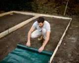 Hombre desplegando tela verde sobre el terreno preparado para una solera de hormigón.