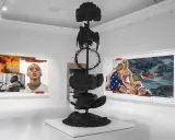 Ein junger Künstler präsentiert seine Werke: ein Porträt vor Flammen, eine Skulptur mit Dornenkrone und ein Bild zweier Männer am Fluss.
