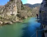 Persona camina por pasarela metálica junto a río turquesa, explorando las rutas de Burgos. Paisaje rocoso y vegetación de ribera.
