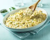 Pyszne, kremowe mac n cheese z ziołami, podane w białej misce. Drewniana łyżka nabiera porcję ciągnącego się sera.