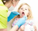 Mała dziewczynka z otwartymi ustami podczas pierwszej wizyty u dentysty. Dentystka w niebieskich rękawiczkach bada jej ząbki.