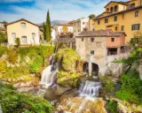 Cascata e mulino in pietra, tra i borghi della Toscana. Case gialle e verdi colline sullo sfondo.