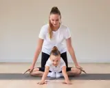 Frau und Kind machen Yoga. Das Kind krabbelt auf der Matte, die Frau unterstützt es. Yoga mit Kindern unter 3 Jahren fördert spielerisch die Bewegung.