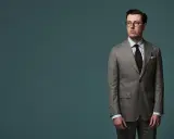 A man in a pinstripe suit, with a brown tie and glasses, stands against a teal background. The jacket length appears to be ideal, hitting just below the seat.