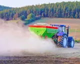 Traktor rozsiewa wapno do ziemi, tworząc chmurę pyłu na polu. W tle las i wzgórza.