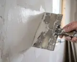 A hand uses a trowel to apply white spackle to a textured wall.