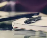 Czarny długopis spoczywa na otwartym notatniku. Obok leży laptop, gotowy do pracy. FGŚP.