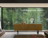 Madia moderna con ante a righe diagonali, un tavolino basso con oggetti decorativi e una grande vetrata su una foresta lussureggiante.