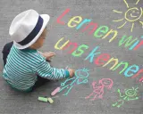 Kleiner Junge mit Hut malt mit Kreide auf Asphalt. Namensspiele für Kinder: "Lernen wir uns kennen!" mit Sonne und Figuren.
