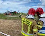 Strażacy w akcji. W tle lądujący śmigłowiec, który wspiera pracę w straży pożarnej.