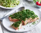 En saftig bit confiterad lax, generöst toppad med färska örter, serveras på en vit tallrik. Bredvid syns en fräsch sallad och några röda rädisor.