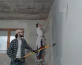 Para z zaangażowaniem wykonuje poprawki na pomalowanej ścianie. On trzyma wałek, ona stoi na drabinie.