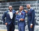 Three groomsmen in stylish navy blue suit combinations, featuring burgundy bow ties and pocket squares, pose in front of a waterfall.