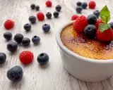 Creme brulee mit karamellisierter Kruste, garniert mit frischen Himbeeren und Blaubeeren, umgeben von weiteren Beeren auf hellem Holz.