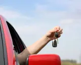 Ręka trzyma kluczyki do samochodu, symbolizujące umowę użyczenia. Czerwony samochód i błękitne niebo w tle.