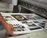 Mani che sistemano fogli stampati su una macchina da taglio, parte della post produzione di una rivista.