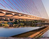 El puente de Castilla-La Mancha se refleja en el río, con sus cables rojos y el cielo azul.