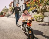 Un bambino con un mantello da supereroe prova ad andare in bicicletta, mentre suo padre corre dietro per aiutarlo. Mio figlio non riesce a pedalare, ma si diverte un mondo!