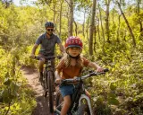 En flicka och en man cyklar på en stig i en lummig skog. Solen lyser genom träden och skapar ett vackert ljusspel.