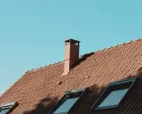 Ceglany dach z kominem i oknami dachowymi. Woda w kominie po deszczu powoli paruje w słońcu.