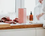 Różowe pudełko z kolagenem B. ESTÉE, szklanka wody i ręcznik. To estetyczne zdjecia produktowe.
