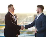 Dwóch mężczyzn w garniturach ściska sobie dłonie na tle miasta.