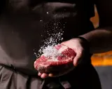 Dłoń kucharza doprawia solą soczysty kawałek mięsa, przygotowując go do grillowania. To początek drogi do idealnych rodzajów steków.