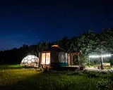 Nocne niebo nad Jurą Krakowsko-Częstochowską. Dwa noclegowe domki, jurt i kopuła, oświetlone ciepłym światłem, otoczone zielenią.