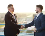 Dwóch mężczyzn w garniturach ściska sobie dłonie na tle miasta.