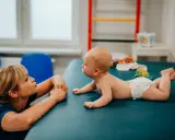 Terapeuta zachęca 3 miesięczne dziecko, które nie chce leżeć na brzuchu, do ćwiczeń na macie.