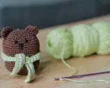 Os **sonajeros de crochet patrones gratis** son adorables. Un osito marrón con bufanda verde y blanca, junto a ovillos de lana y una aguja de crochet.