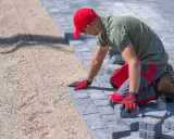 Pracownik w czerwonej czapce układa kostkę bauma na podsypce.