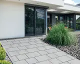 Nowoczesna ścieżka z szarych płyt betonowych zamiast kostki, prowadząca do minimalistycznego domu z dużymi oknami.