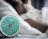 Ein Wecker neben einer schlafenden Person. Die Zeit ist kurz vor 2 Uhr. Ein Symbol für den zirkadianen Rhythmus.