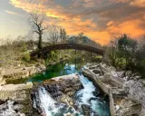 Un puente arqueado cruza un río verde, con cascadas y ruinas de piedra. Un paisaje idílico que invita a descubrir pueblos bonitos cerca de Puente Viesgo.