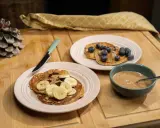 Pancakes ig bas farine complète, garnis de bananes, de chocolat et de myrtilles, avec un bol de beurre d'amande.