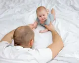 Tata pomaga dwumiesięcznemu dziecku obrócić się z brzucha na plecy. Maluch z zaciekawieniem patrzy na tatę.