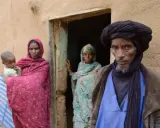 Ein Tuareg-Mann mit schwarzem Turban und ein Kind mit seiner Mutter vor einem Lehmhaus in der Sahara.