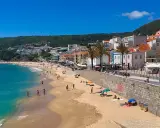 Sonne, Strand und Meer in Sesimbra, Portugal. Menschen genießen den Tag am goldenen Sandstrand, während im Hintergrund die Stadt und grüne Hügel aufragen.