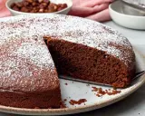 Ein saftiger Nutella Kuchen, bestäubt mit Puderzucker, wartet darauf, angeschnitten zu werden. Daneben eine Schüssel mit Haselnüssen.