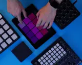 Des mains jouent sur une tablette avec des pads colorés, entourée de contrôleurs pour créer de la musique. Un sampler sans ordinateur en action.